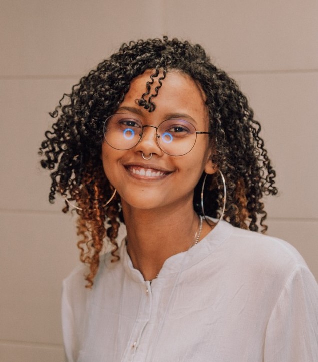 foto de Maria Eduarda, estou de óculos, camisa branca e tenho cabelo cacheado na altura dos ombros